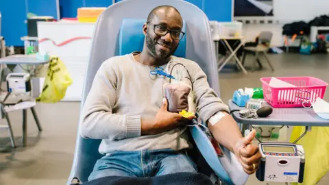 A man wearing a beige top and blue jeans with his sleeves rolled up as he is donates blood. he is holding a bag of blood with wires coming in and out of his arm. 