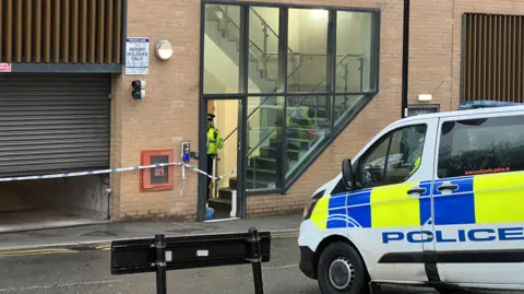 The outside of an apartment building. Inside the glass door stands a police officer. There is police tape across the entrance to the underground garage, to the left of the tenants' entrance. A police van sits on the road outside.