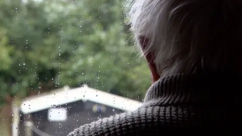 The back of an older man with white hair. He is looking out of a window into a garden with a black shed with a white roof. There is rain on the window