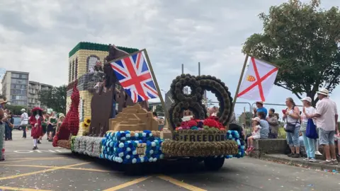 A float celebrating Liberation 80. At the front are huge numbers: 80. Under them it reads FREEDOM. There is a Jersey flag and a Union Jack flag flying. Behind this is a large green and white house and a girl is standing on a balcony on it. Behind the float is a man dressed in a red Charles I style costume complete with a big hat. Spectators are watching the float.