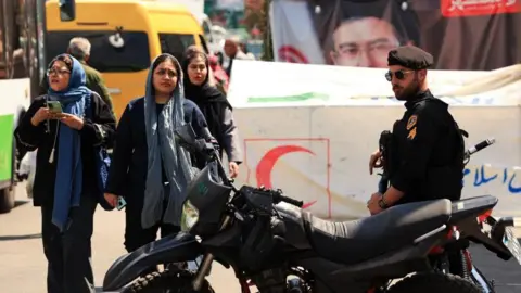 Iranian women walk past a police officer standing guard on a street, amid a ceasefire between the U.S. and Iran, at Tajrish Square in Tehran, Iran