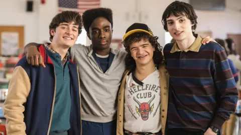 Netflix Noah Schnapp in character as Will Byers alongside Finn Wolfhard as Mike Wheeler, Gaten Matarazzo as Dustin Henderson and Caleb McLaughlin as Lucas Sinclair. The group have their arms wrapped around each other and they are smiling at the camera. 