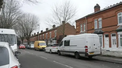 A residential street lined with terraced houses and trees. Cars and vans are parked both sides of the road.