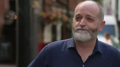 Middle aged man with receding hair and a grey beard wearing a blue shirt stands in front of a pub with colourful hanging baskets