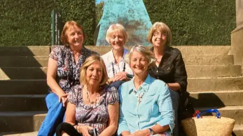 Caroline Velvick Five women sit on steps. Three women on the back row and two in the front. They are wearing jeans and blouses and one has glasses. 