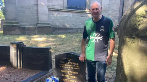 Lionel Bird in green and black football tshirt and dark jeans in standing in a cemetery next to a black gravestone with the words, in memory of Willie Stanley