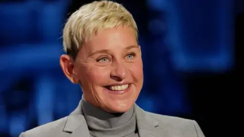 Getty Images Ellen DeGeneres smiling, wearing a grey turtleneck apical  and grey jacket