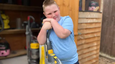 A boy with short blond hair is stood outside of a garden shed with his hands on top of a yellow and grey vacuum cleaner. The boy smiles at the camera and is wearing a light blue T-shirt and navy blue jogging bottoms.