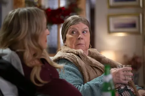 BBC Studios Scotland / Graeme Hunter A still from the Christmas Special shows Christine (Elaine C Smith) turning to Michelle (Joy McAvoy) as they sit on a couch. Christing is holding a glass of what looks like whisky.