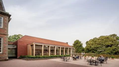 Greg Holmes An outdoor seating area with black tables and chairs. There is a red brick building to the left that has a glass front and wooden pillars that divide the windows.
