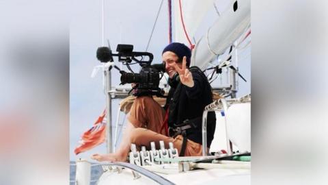 Eleanor Church sitting on a boat while filming the ocean and giving a peace sign