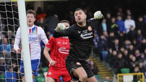 Pacemaker Aaron Hogg is kicking the ball in a football match. He is wearing an all black goalkeeper kit with gloves. Two other male players are in the background - one is running towards him from the left.