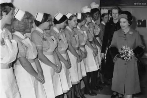 NHS Humber Health Partnership Queen Elizabeth II holds a bouquet of flowers as she walks past a line of student nurses, all wearing matching nurse uniforms and hats. The photo is in black and white.