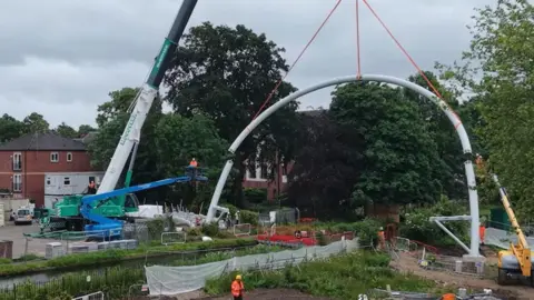 Erewash Borough Council A white curved structure being lifted into place over a canal by several cranes.