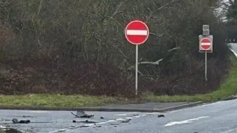 A dual carraigeway is surrounded by trees. A no entry sign can be seen. A car with its lights on is in the distance. 