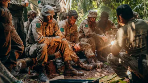 British Army Soldiers in camouflage uniforms sitting in a clearing. They have dirt on their faces. Another soldier is kneeling in front of them, teaching them jungle skills. 