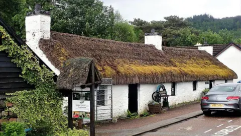 The Glencoe Folk Museum which is a single-storey white building with a thatched roof with a car parked in front of it and trees behind it