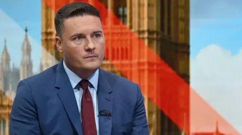 Wes Streeting, wearing a blue suit, white shirt, and red tie, sits in a red chair on the set of a Sunday with Laura Kuenssberg. 