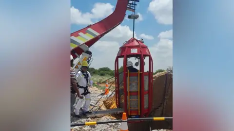 Giwusa A man in a white boiler stands next to a crane that is about to lower a cage into a mine shaft.