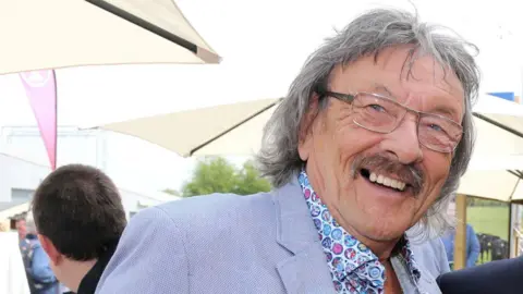 Big T has long grey hair and glasses. He is wearing a light-blue blazer and multi-coloured shirt.  He is smiling, behind him are cream umbrellas.