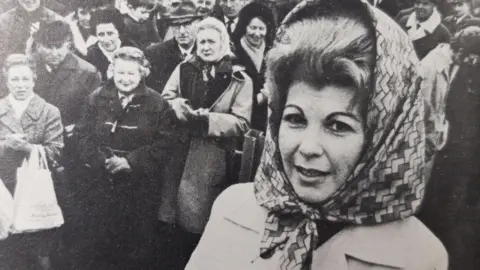 Black and white photo of Sally Oppenheim-Barnes stands in front of a crowd in the mid-20th century, wearing a patterned covering over her big hair.