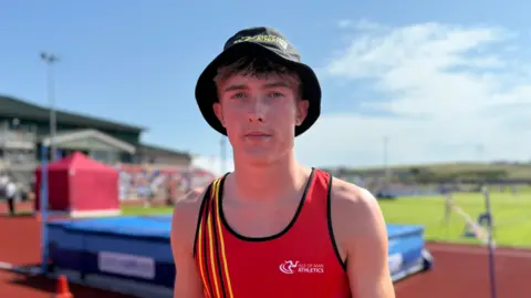 Regan Corrin wars a black bucket hat and a red vest. He stands near the high jump equipment of a bar and a blue crash mat by an orange race track.