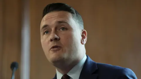 A head and shoulders shot of Wes Streeting mid-speech. He is wearing a navy suit, a white shirt and a navy and red striped tie. 