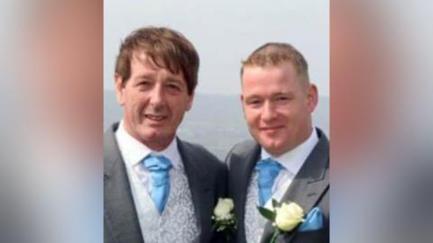 A father and son with their arms around each other, they are wearing wedding suits and boutonnieres
