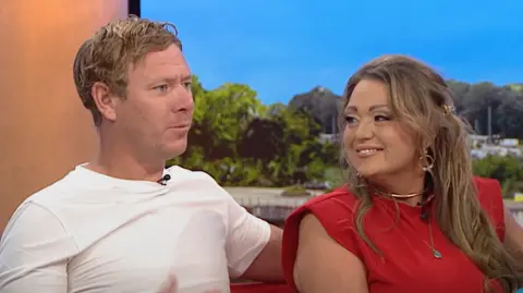 A man with short blonde hair and a white T-shirt is sitting on the BBC Breakfast sofa next to a woman with long dark blonde hair and a red top. He is talking and she is smiling.