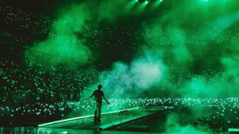 Oliver Akinfeleye Wizkid on stage at The 02 arena in London in November 2021. He has his back to the camera and is facing the audience, whose phones light up the eerie green atmosphere as dry ice lingers in the air.