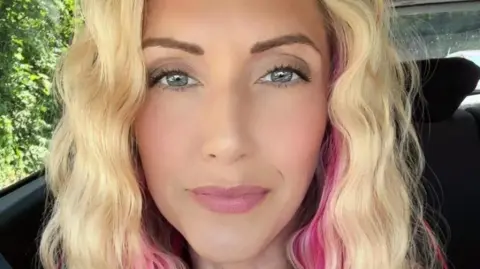 Jessica, who has curly blonde hair and pink highlights, with blue eyes, smiles as she sits in a car