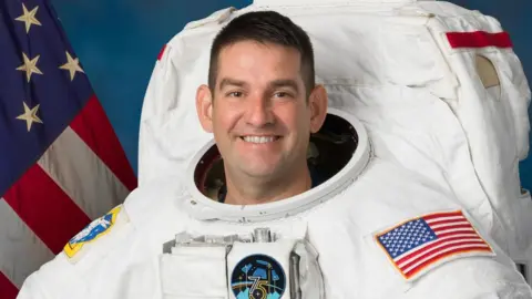 NASA A man wearing an astronauts uniform with an American flag on the arm is smiling