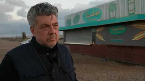 A middle-aged man with dark greying hair stands on a pebble beach outside his green fast food kiosk