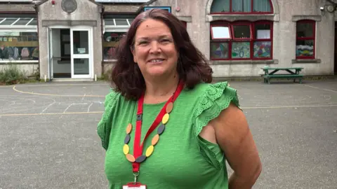 A woman with dark hair down to her shoulders wearing a green top and multicoloured necklace standing in a school playground with a school building behind her. 
