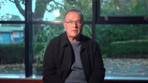 A man with short grey hair and silver glasses is facing the camera. He is wearing a grey jumper and a black jacket. He is sitting in front of large windows, behind which are a tree and hedges