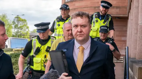 Jordan Linden, wearing a dark blue suit, pink shirt and yellow tie, leaves court as reporters ask questions. There are four police officers on the steps behind him, and a man in a yellow polo shirt.