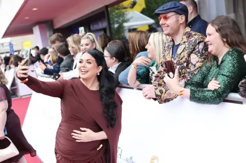 Getty Images Scarlett Moffatt holding her stomach in one hand and a phone in the other, taking a selfie with some fans behind a barrier