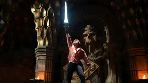 PA Media Film still from Masters Of The Universe. Nicholas Galitzine as Adam holding up a bright blue sword inside a dark castle setting, with flaming roches. 