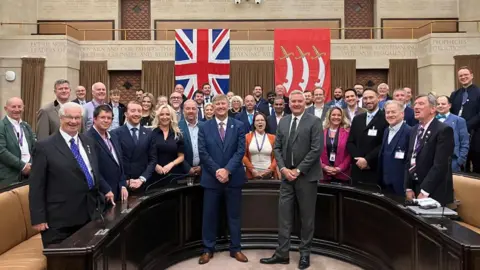 Reform UK A group of councillors standing in a semi-circle in a council chamber.