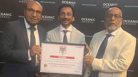 Basmati restaurant Three men wearing glasses, a shirt, a tie and a jacket are holding a framed certificate that includes the words outstanding restaurant of the year Basmati restaurant. They are in front of a black board including the words Oceanic Awards. 