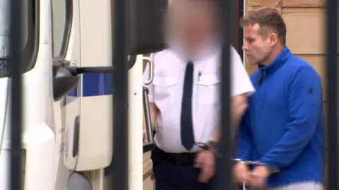 Rory Trainor, in a blue fleece, is handcuffed to a prison officer. The prison officer is wearing a white shirt, black trousers and black tie. Trainor has brown hair. The prison officer's face is blurred. There is a white van to the left.