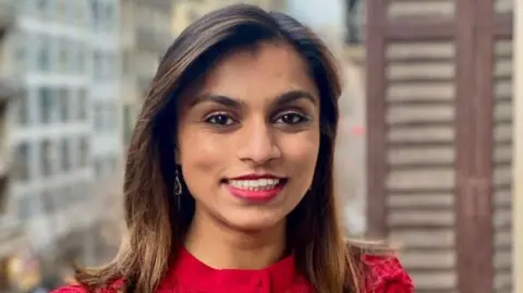 Tanuvi Ethunandan is pictured above her shoulders. She has straight, dark brown shoulder-length hair. She has brown eyes and is wearing red lipstick. She is also wearing a red top. She is pictured in front of a city landscape.