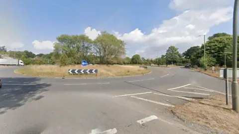 A Google Maps image of a roundabout. There is a lorry to the left of the road and a car approaching from the right. 