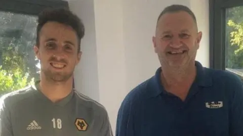 Diogo Jota is on the left in a grey Wolverhampton Wanderers football shirt, and on the right is Stephen Billingsley, wearing a blue polo shirt. Behind them is a white wall with two windows in it
