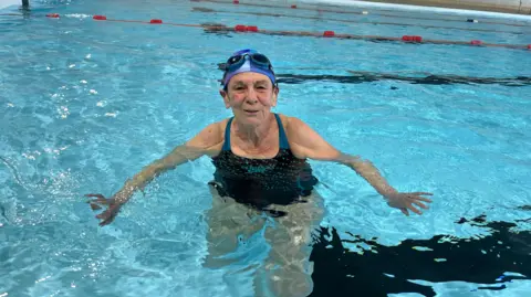 Hilary Butler, 76 is in a public swimming pool alone, she is wearing a black and blue costume and a blue swimming cap with swimming goggles on top of her head.