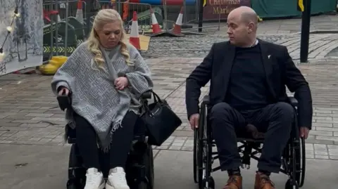The Wheel Life Liverpool Lisa McCann has long blonde hair and is wearing a grey shawl and black trousers and white trainers. She is joined by Liam Robinson who is wearing a black suit, black jumper and brown shoes. They are both using wheelchairs on a Liverpool City Centre path.