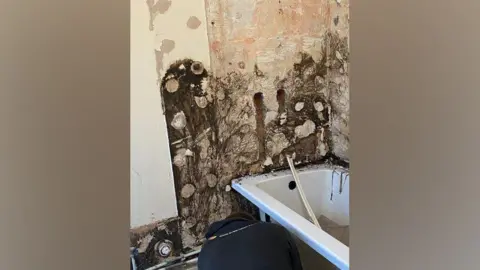 Tiles have been removed in a bathroom revealing dark mould on the wall behind a white bathtub