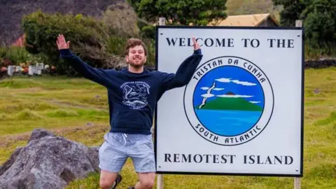 Jake Rosmarin Jake Rosmarin smiles beside a sign that says "Welcome to the remotest island, Tristan da Cunha, South Atlantic".