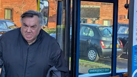 Brian Wernham Alan Mitchell, with short grey hair, leans on a mobility aid, standing next to an open bus door. A row of houses can be seen behind.
