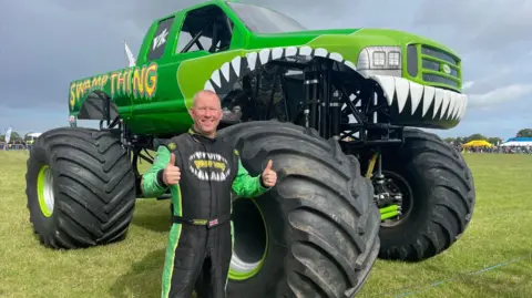 Tony Dixon smiles with his thumbs up while stood in front of a green monster truck with a mouth and the words "Swamp Thing" spray painted on it drives through a field as people behind a barrier watch on. 